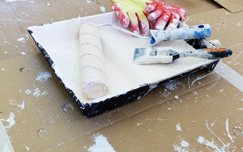 Paint roller and brush resting in a paint tray on a cardboard drop cloth