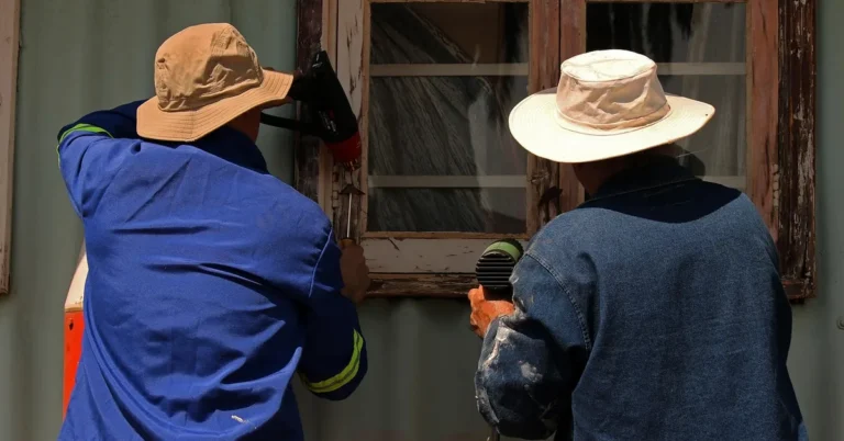 Two workers stripping old paint from a wood window frame during restoration