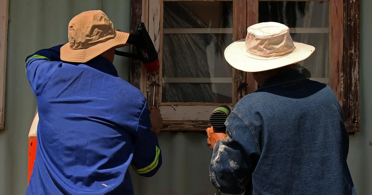 Two workers stripping old paint from a wood window frame during restoration