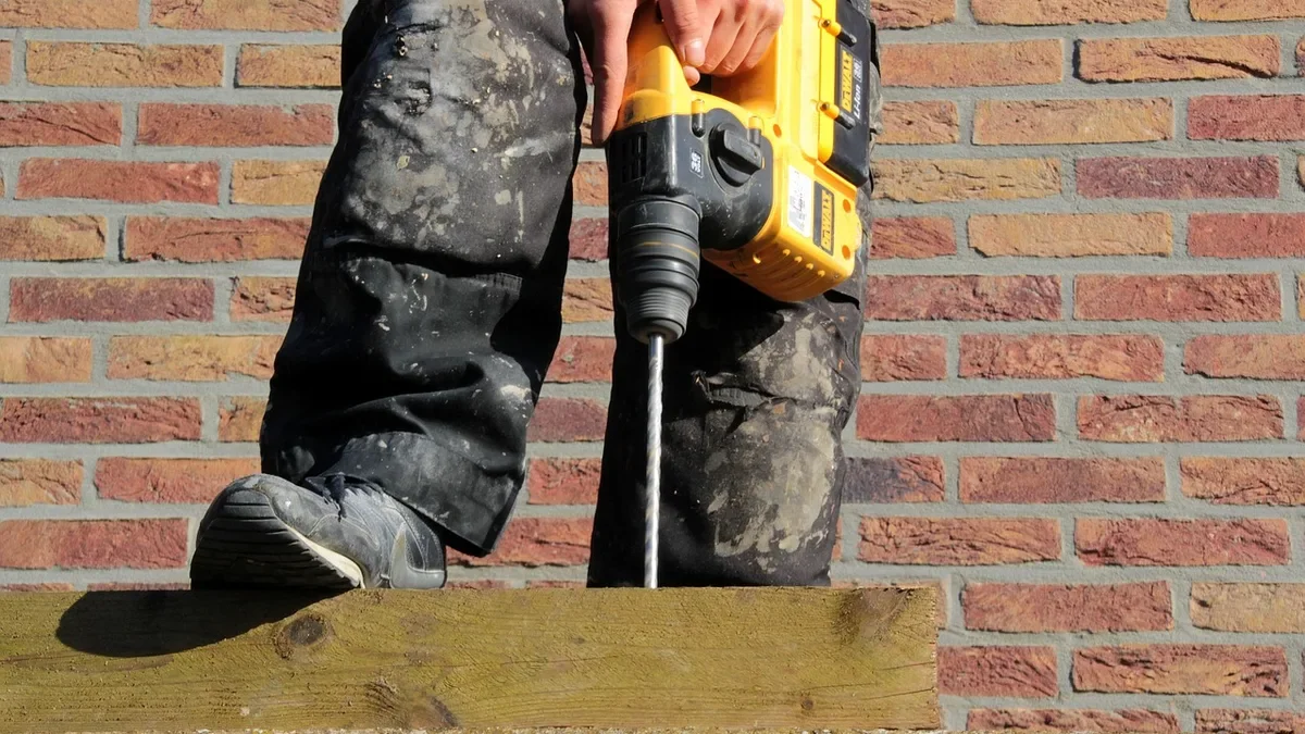 Worker holding a hammer drill with a masonry bit beside a wood plate on a brick wall ledge