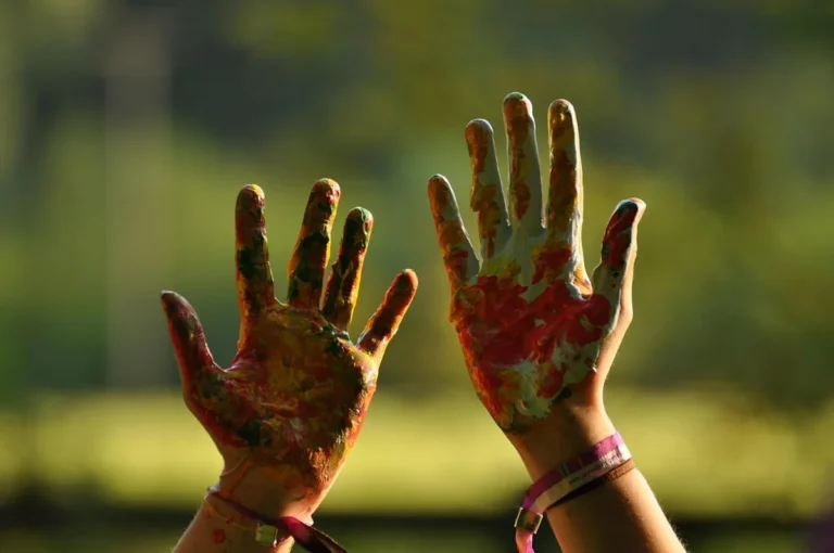 Two hands covered in dried multicolored paint held up against a green background