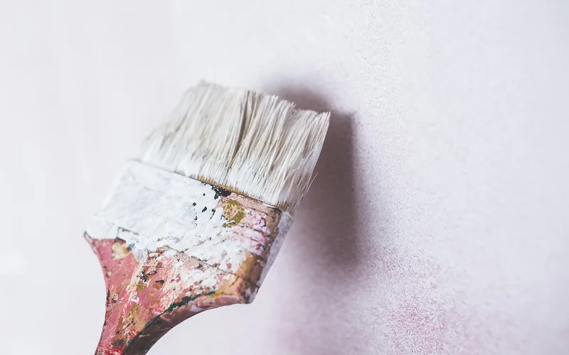 Close-up of a paint brush applying fresh paint along a wall edge near masked trim