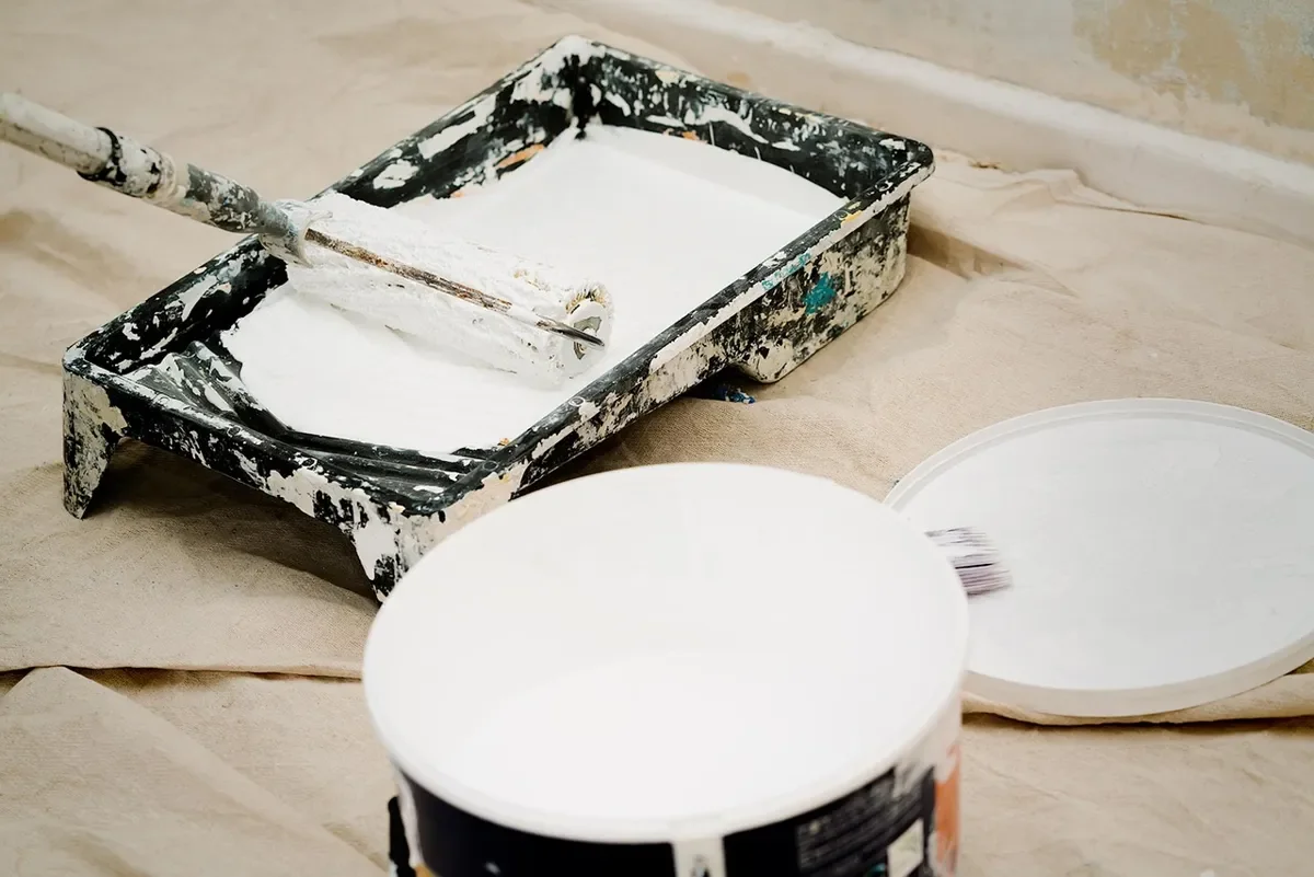 paint roller resting in a white-stained paint tray on a drop cloth beside an open paint can