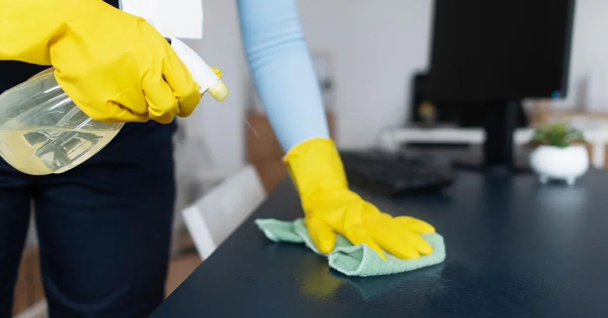 Gloved hand wiping a laminate desk surface with a damp microfiber cloth and spray bottle