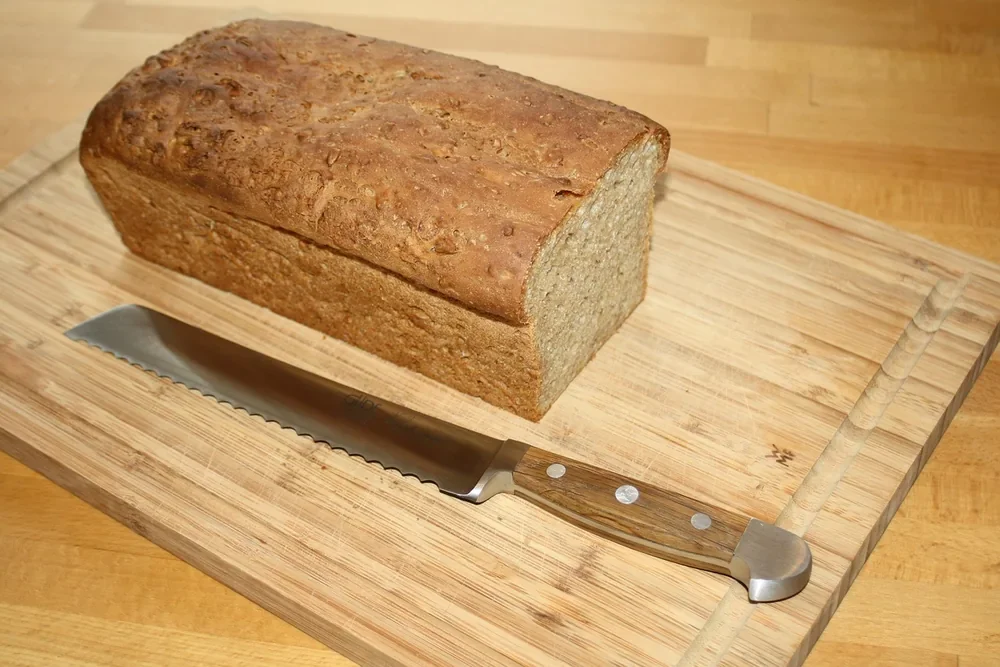 Bread knife with a dark wooden handle resting on a wooden cutting board next to a loaf