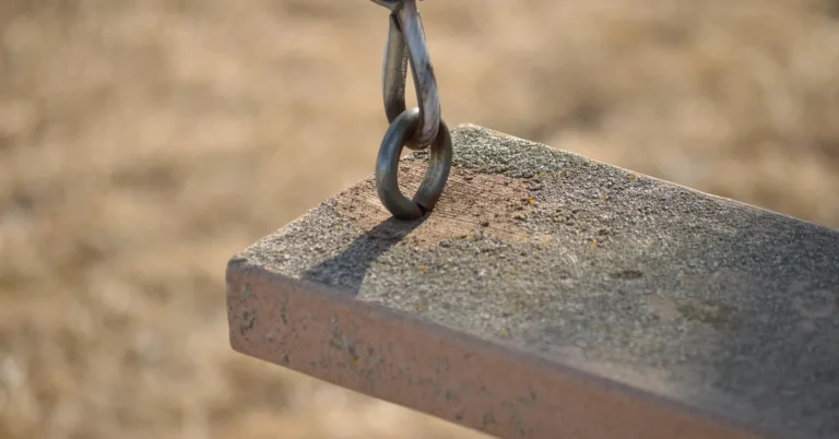 Close-up of a weathered wooden swing plank with a metal chain hook, showing the two swing set materials side by side