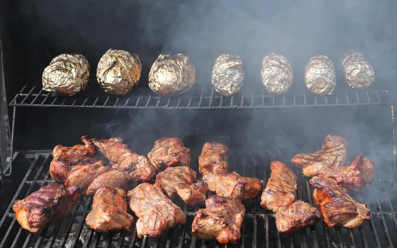Meat and foil-wrapped potatoes cooking on a two-level smoker rack with smoke