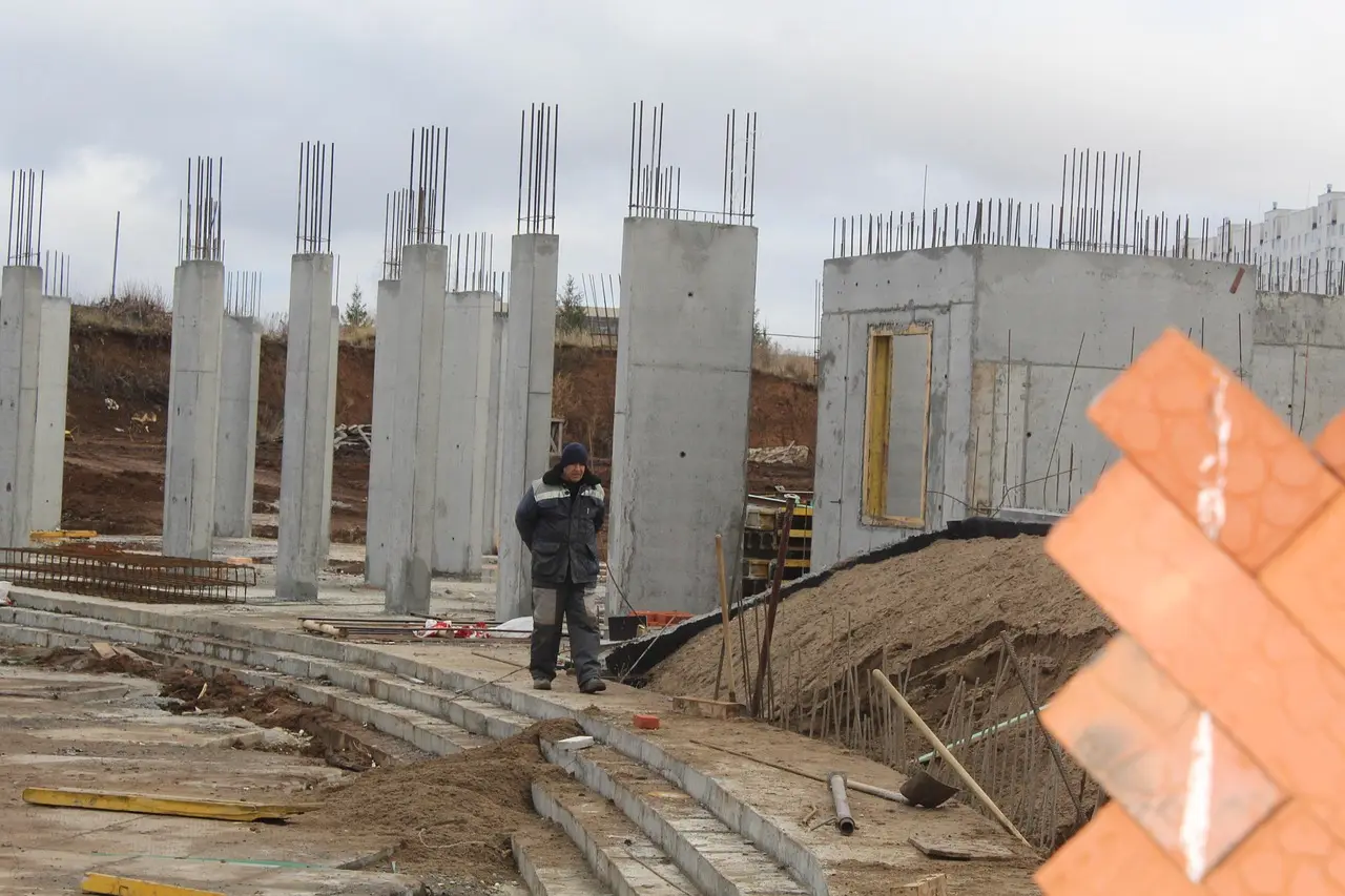 Contractor measures footing depth beside an excavated trench with damp soil and frost on the ground